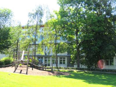 Blick auf die Grundschule. Bäume und ein Klettergerüste stehen im Pausenhof der Schule